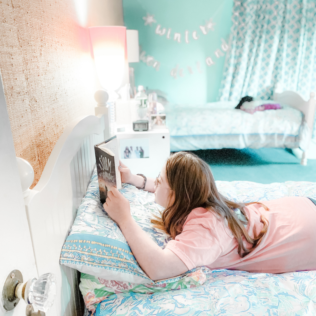 Penny rests on her stomach on her bed reading a book that is propped on a pillow