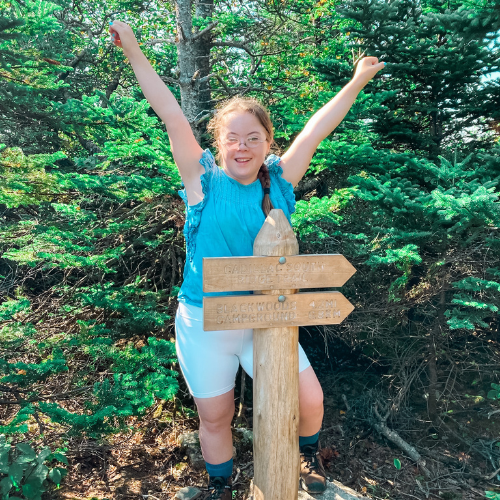 Penny stands in front of a trail sign and and smiles with her arms raised into the air in celebration