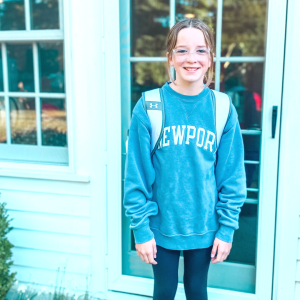 Marilee stands in front of windows of a house and smiles at the camera. She wears a backpack.