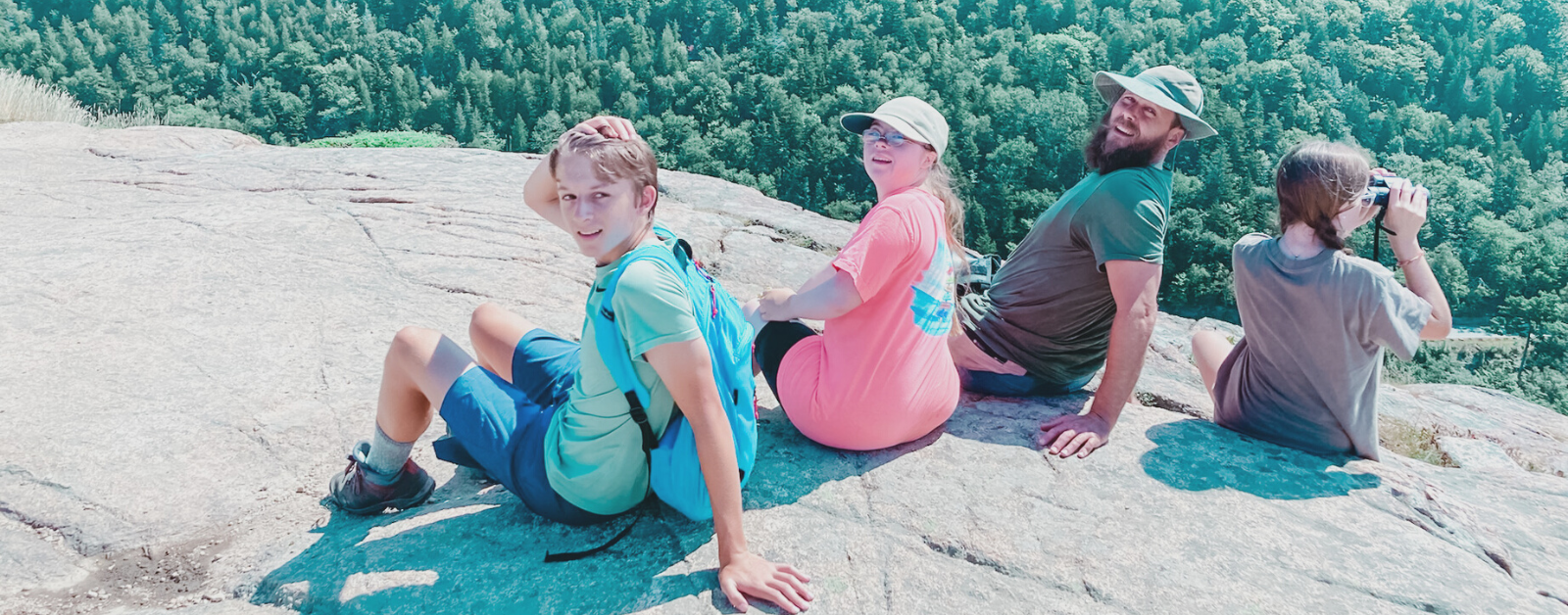 William, Penny, friend, and Marilee sit on a large rock with a forest far below them.