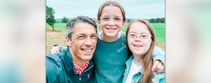 Peter, Marilee, and Penny stand outside with their arms around each. They smile at the camera, and there are green fields and grass behind them.