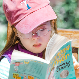 young Penny wears a Taft hat and sits outside on a bench at Taft. She is reading a Beverly Cleary book