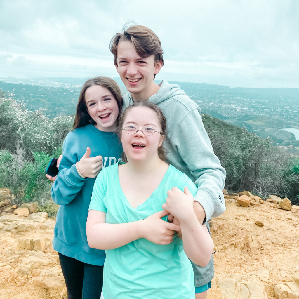 Marilee, William, and Penny smile at the camera with their arms around each other. They stand on an outcropping with trees and a city below them.