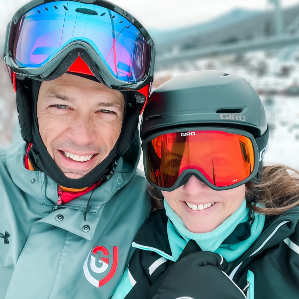 Peter and Amy Julia smile for a selfie. They are wearing ski goggles and there is a snowy slope behind them