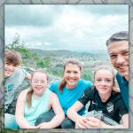 William, Penny, Amy Julia, Marilee, and Peter smile for a selfie on a rock outcropping overlooking a valley