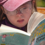 photo of young Penny wearing a pink hat, sitting on a bench outside, and reading a book