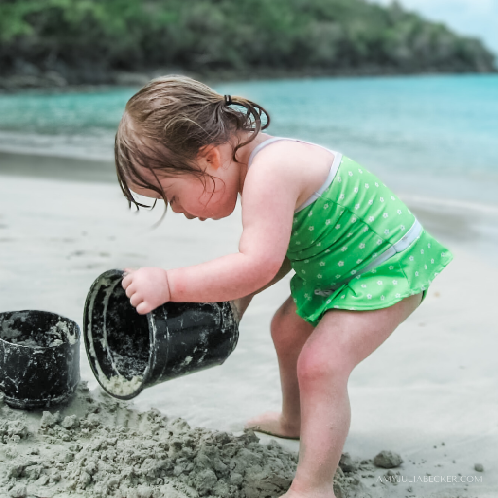 toddler Penny dumping a bucket of sand at the beach