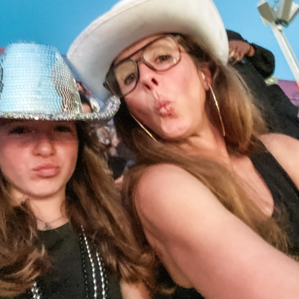 Marilee and Elizabeth wear cowboy hats and smile for a selfie as they sit in stadium seating