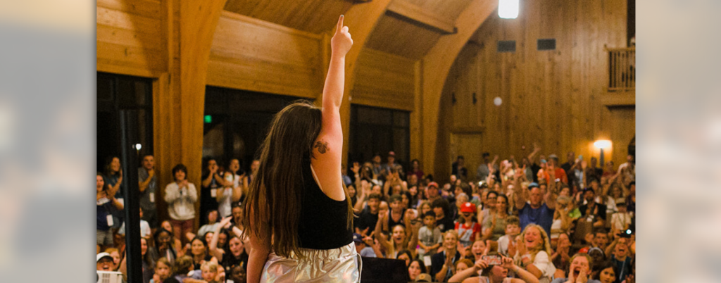 Penny stands with her back to the camera on stage at the Hope Heals talent show with her arm raised in the air and facing an excited audience