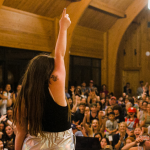 Penny stands with her back to the camera on stage at the Hope Heals talent show with her arm raised in the air and facing an excited audience
