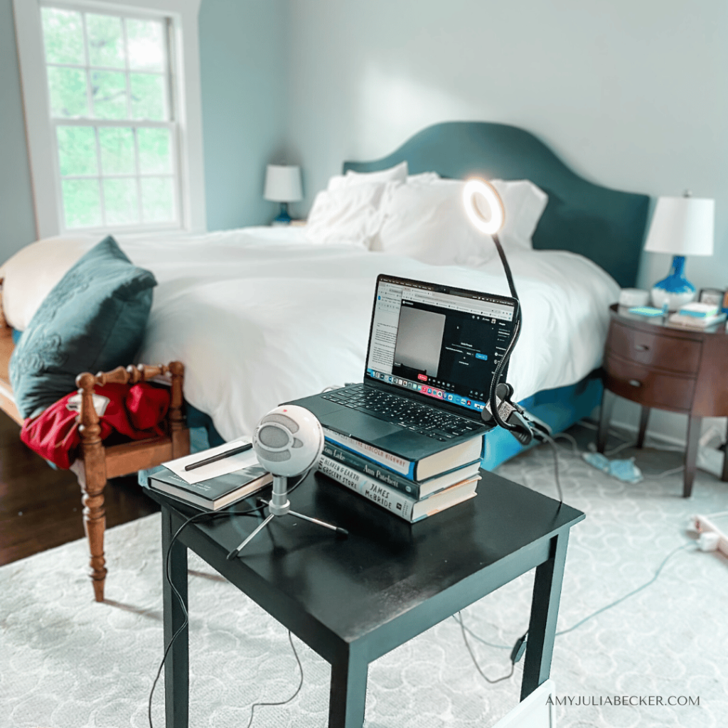 a photo of a desk with a laptop on top of a stack of books, a lamp, and a mic in a bedroom with the bed in the background