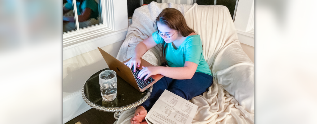 photo of Penny sitting inside on a comfy white chair next to large windows; she is typing on a laptop