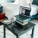a photo of a desk with a laptop on top of a stack of books, a lamp, and a mic in a bedroom with the bed in the background