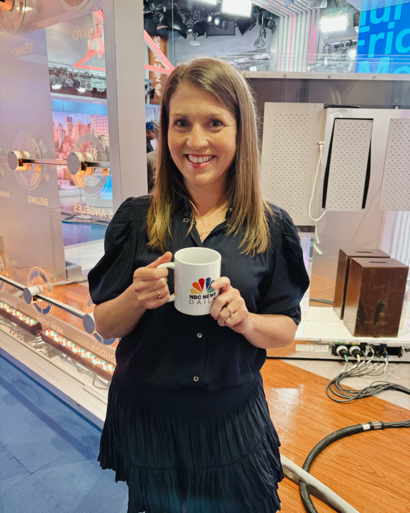 Amy Julia stands inside NBC studios and smiles at the camera as she holds an NBC mug