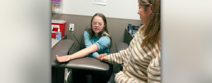 photo of Penny sitting in a chair in a doctor's office. She is waiting to get her blood drawn and Amy Julia is standing by her holding her hand.