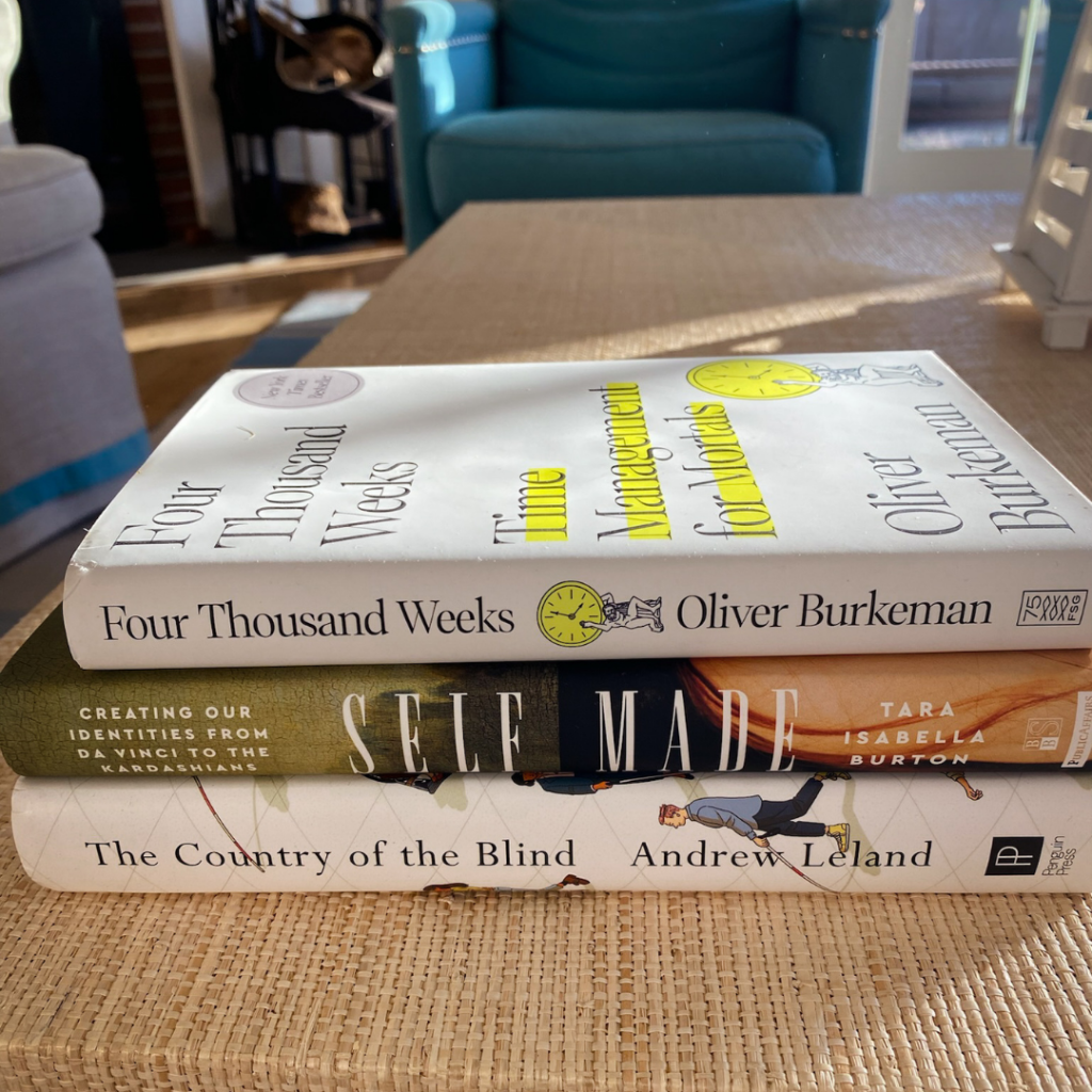 stack of three books (Four Thousand Weeks, Self Made, and The Country of the Blind) on a table with a living room in the background