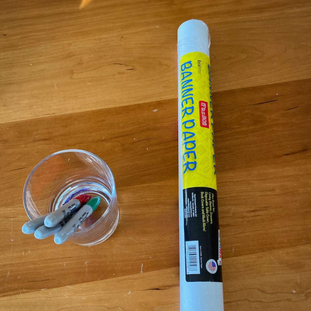 glass cup holding sharpies on a table next to a roll of banner paper