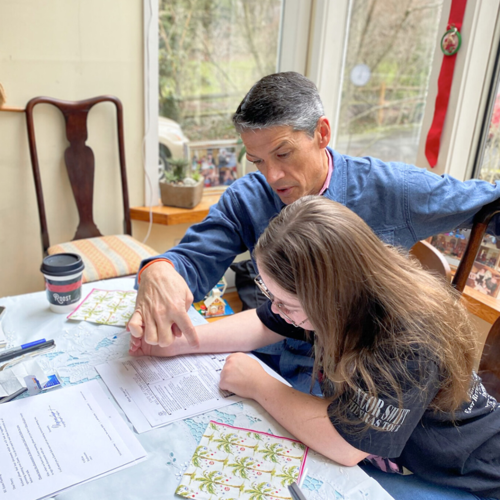 photo of Penny, a young adult with Down syndrome, sitting at a table with her dad. They are looking down at paperwork on the table.