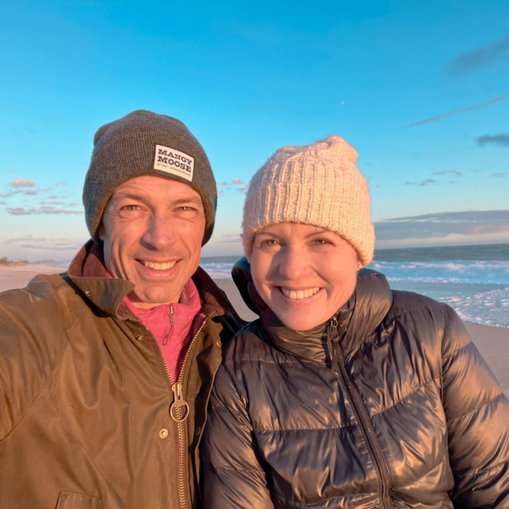 Peter and Amy Julia smile at the camera. They are wearing winter coats and hats with the ocean and blue sky in the background.