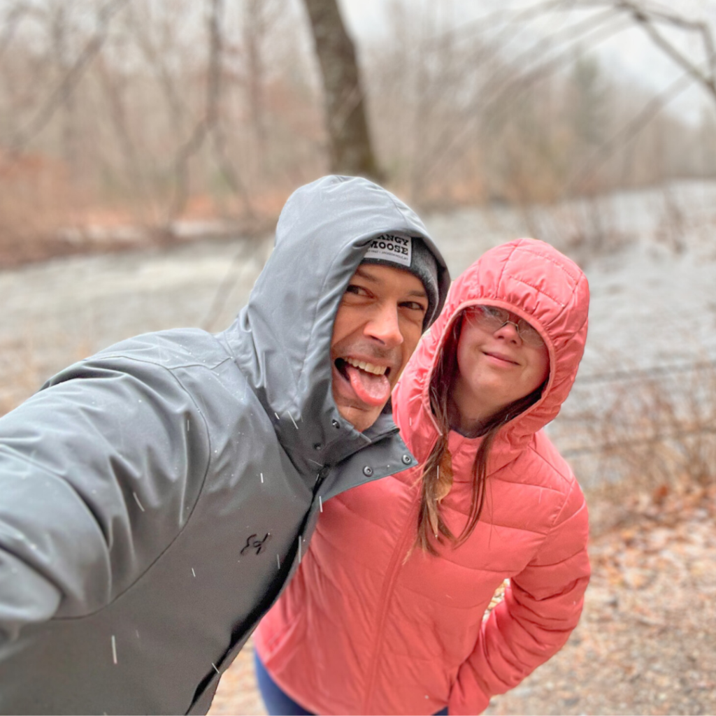 Peter and Penny pose in front of a river. They are wearing winter coats with their hoods up, and Peter is making a silly face at the camera.