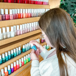 Penny, a young white woman with Down syndrome, stands with her back partially to the camera. She is in front of rows of nail polish and is looking down at a bottle of blue polish that she holds in her hands.