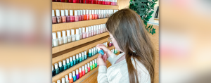 Penny, a young white woman with Down syndrome, stands with her back partially to the camera. She is in front of rows of nail polish and is looking down at a bottle of blue polish that she holds in her hands.