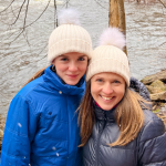 Marilee and Amy Julia smile at the camera. They are wearing winter coats and matching white stocking hats and are standing in front of a river.