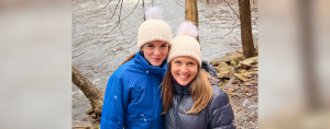 Marilee and Amy Julia smile at the camera. They are wearing winter coats and matching white stocking hats and are standing in front of a river.