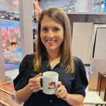 Amy Julia, a white woman wearing a dark blue dress, holds a white mug with the NBC logo. She is smiling at the camera with the NBC studio in the background.