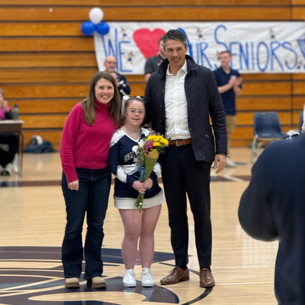 Amy Julia, Penny, and Peter stand at center court and smile towards the camera. Penny is wearing her cheerleading uniform and holding a bouquet of flowers