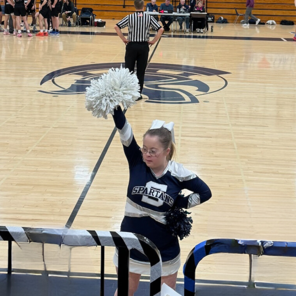 enny, dressed in her blue and white cheerleading uniform, cheers in front of the bleachers with a white pompom held high