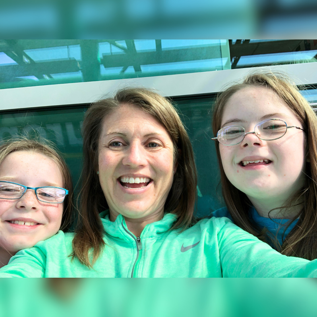 Amy Julia, who is wearing a green jacket, takes a selfie with her two young daughters. They are all smiling at the camera. Marilee is on the left, and Penny is on the right. They are both wearing glasses.