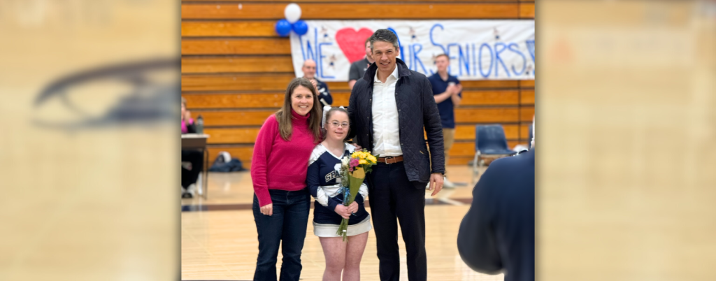 Amy Julia, Penny, and Peter stand at center court and smile towards the camera. Penny is wearing her cheerleading uniform and holding a bouquet of flowers for senior night