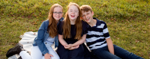 3 children sit on a blanket in the grass and smile at the camera with big grins.