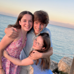 a photo of 3 teen siblings—Marilee wearing a pink dress, William in the middle wearing a blue shirt, and Penny on the right wearing a blue dress—giving each other a group hug in front of a faint sunset over the ocean