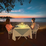 Amy Julia and Marilee sit at a linen-covered table on the beach and look out at the sunset over the ocean