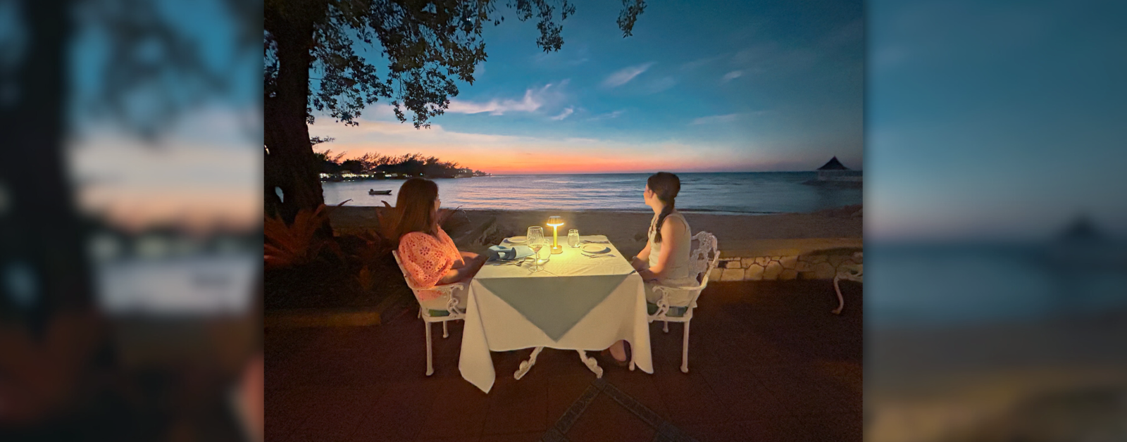 Amy Julia and Marilee sit at a linen-covered table on the beach and look out at the sunset over the ocean