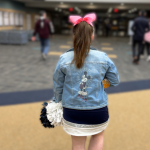 Penny, dressed in her blue and white cheerleading uniform and wearing a jean jacket over it and a pink bow in hair, walks down the school hallway away from the camera