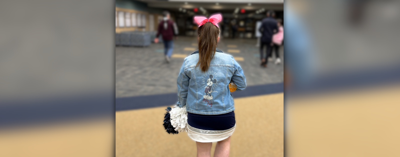 Penny, dressed in her blue and white cheerleading uniform and wearing a jean jacket over it and a pink bow in hair, walks down the school hallway away from the camera