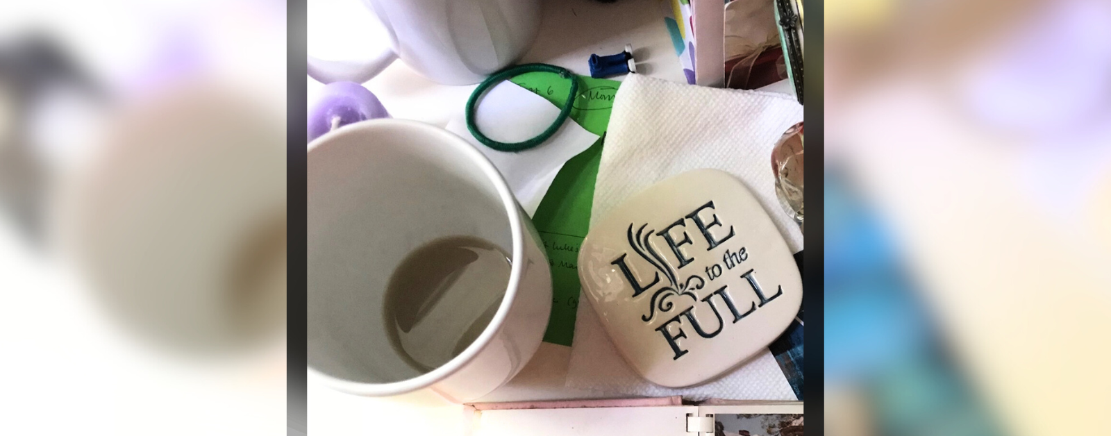 a photo looking down at a cluttered desk; in view is a nearly empty, white mug, a coaster that says "Life to the Full," a hair tie, napkins, and papers and envelopes