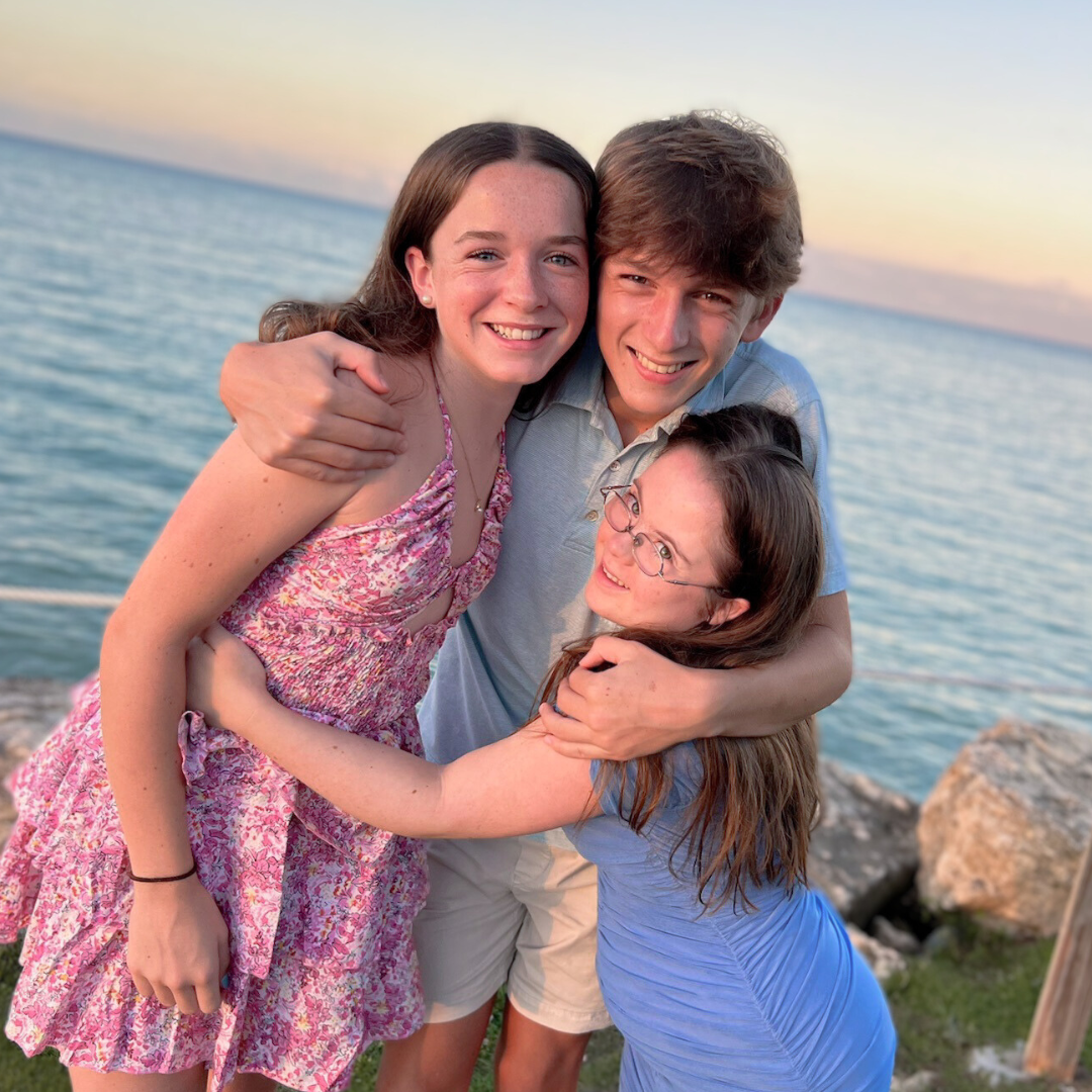 a photo of 3 teen siblings—Marilee wearing a pink dress, William in the middle wearing a blue shirt, and Penny on the right wearing a blue dress—giving each other a group hug in front of a faint sunset over the ocean]