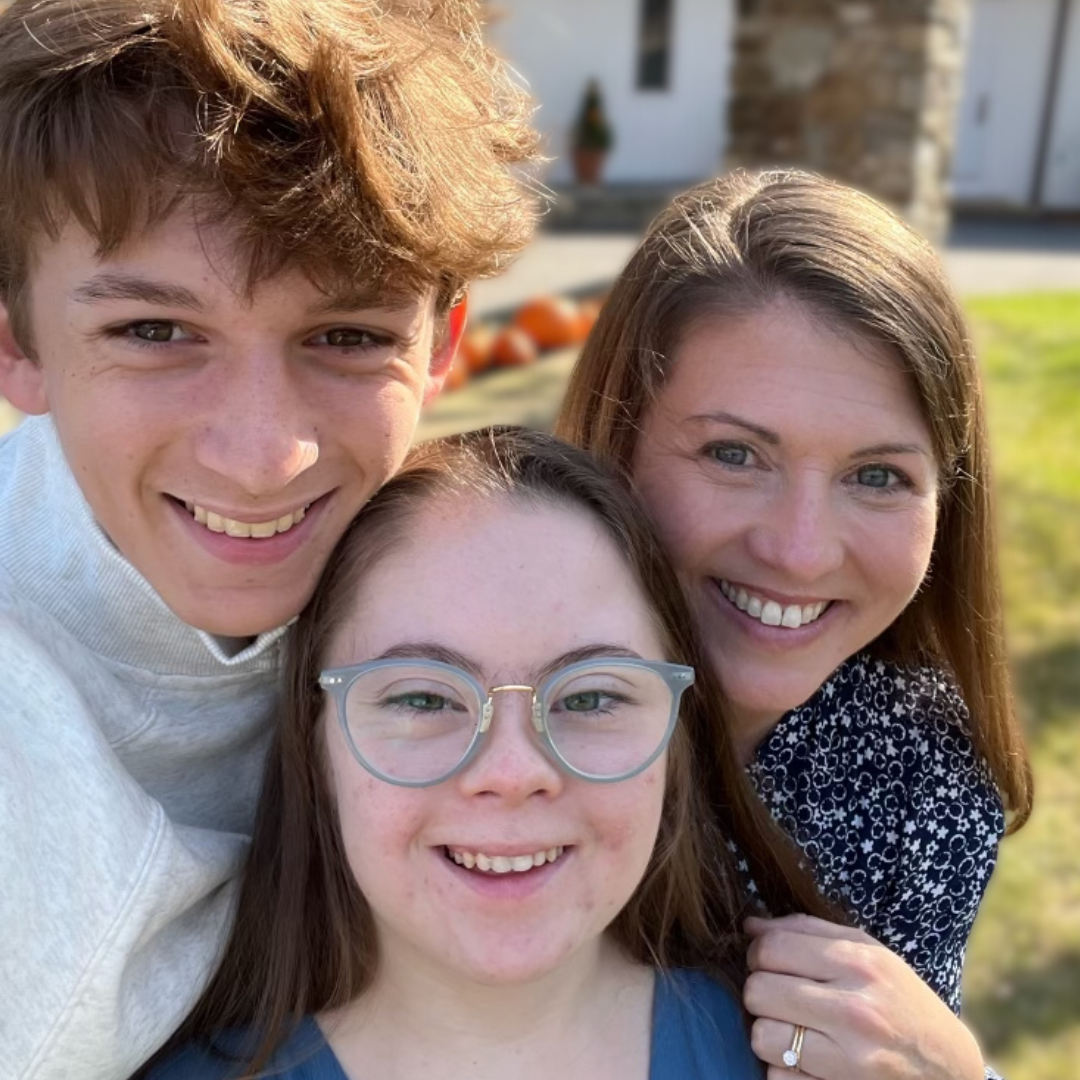 an outdoor photo of William, a teenage boy, Penny, a teenage girl with Down syndrome, and their mom, Amy Julia, leaning in close together and smiling at the camera