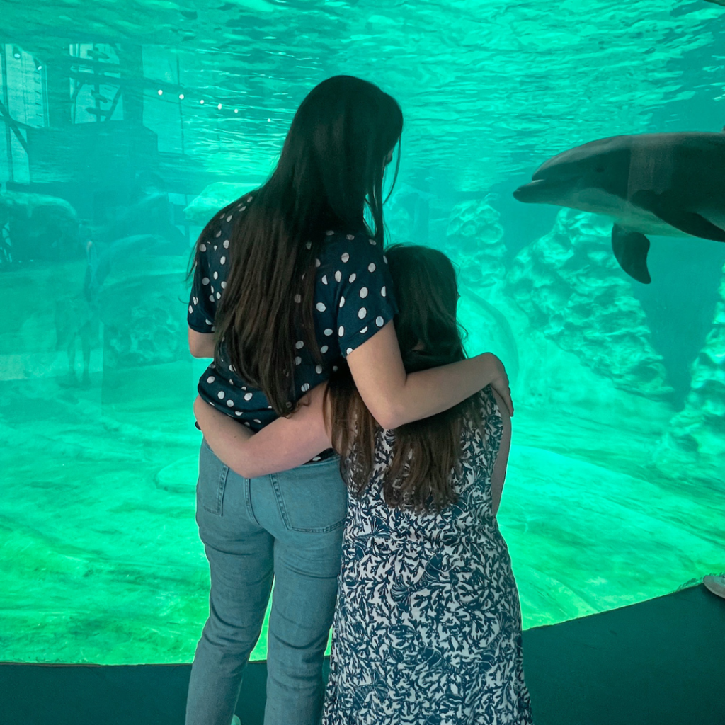 Penny stands next to Alex Wolf, her Hope Heals friend. They are facing away from the camera and looking into an aquarium tank as a dolphin swims toward them