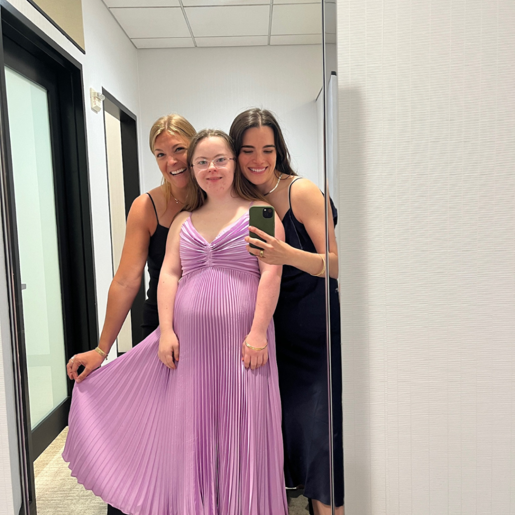 Penny, wearing a pink prom dress, poses in the changing room mirror with two friends from Hope Heals