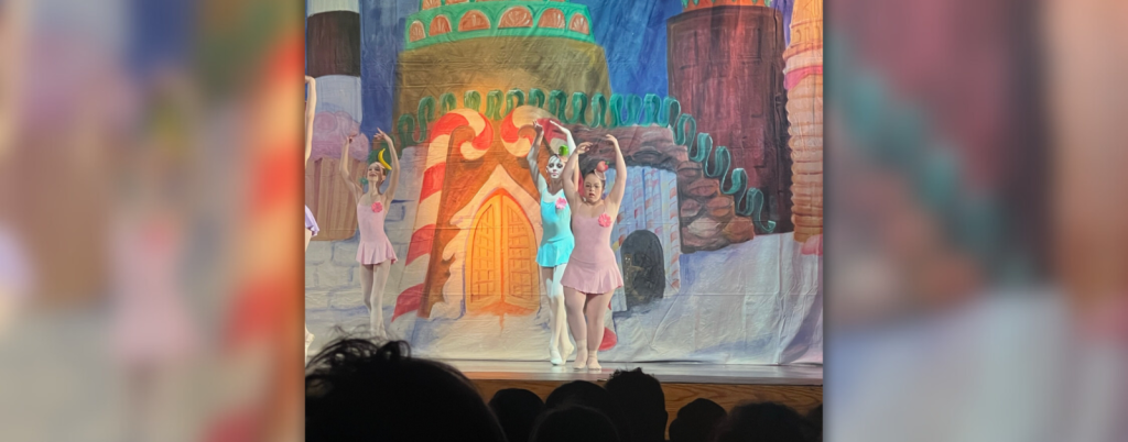 Penny dances on stage with a scene from the Nutcracker behind her. She and two other dances wear tutu dresses and have their arms rounded and raised above their heads