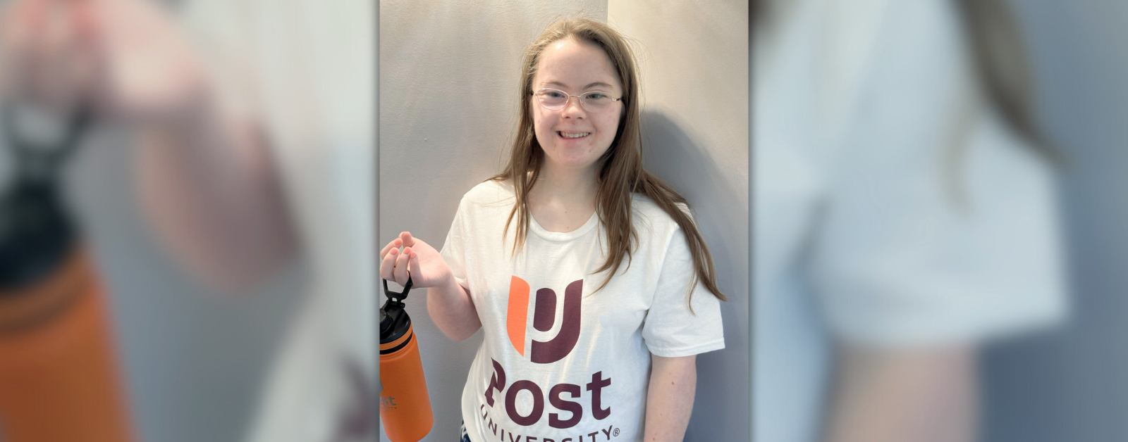 Penny poses inside wearing a Post University t-shirt and holding a Post University orange water bottle as she smiles at the camera