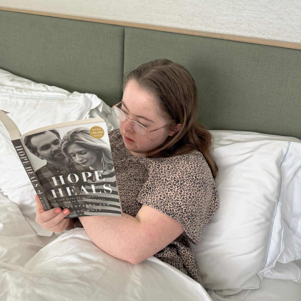 Penny is under the covers at a hotel and is reading the book Hope Heals