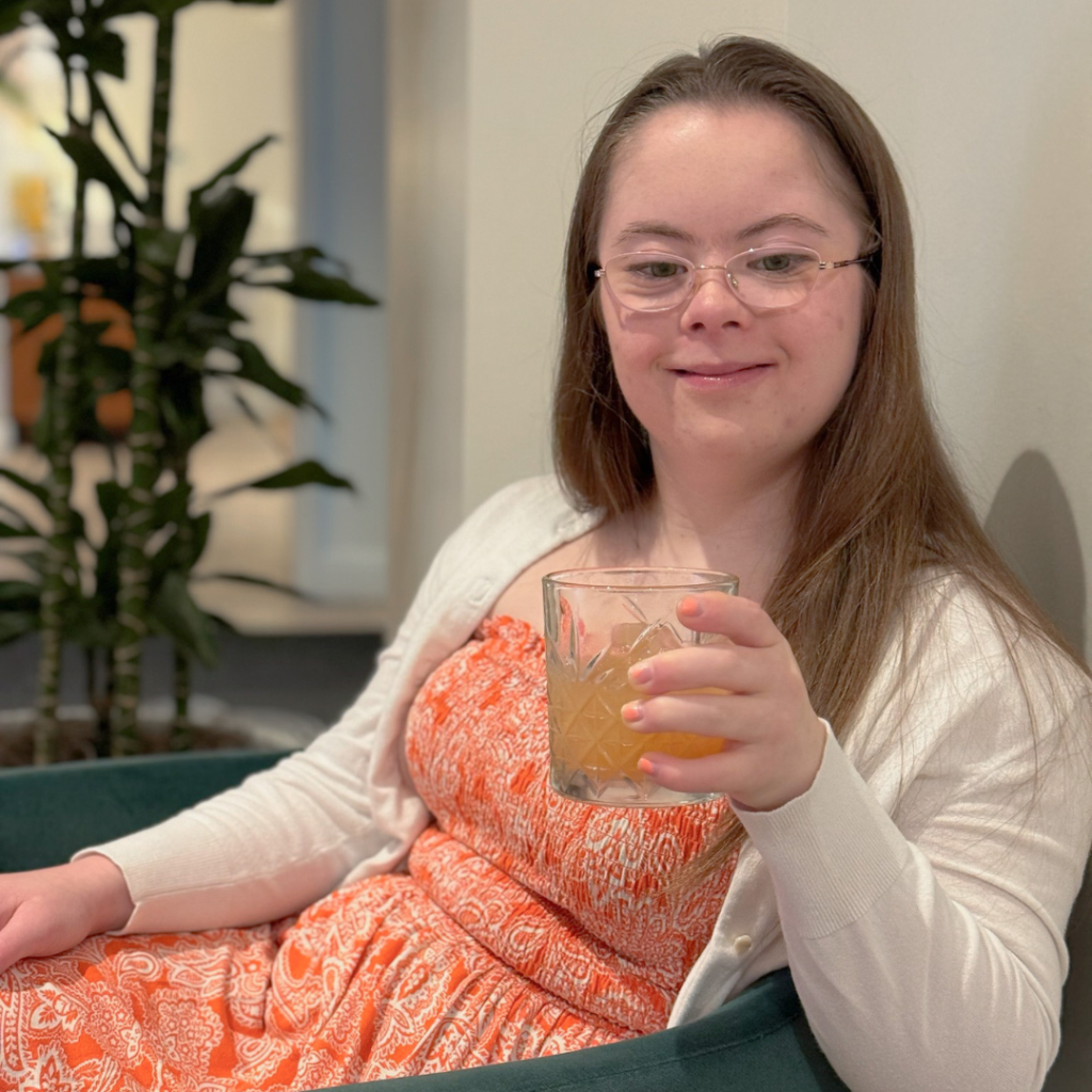 Penny sits in a chair smiling at the camera and holding a drink in her hand
