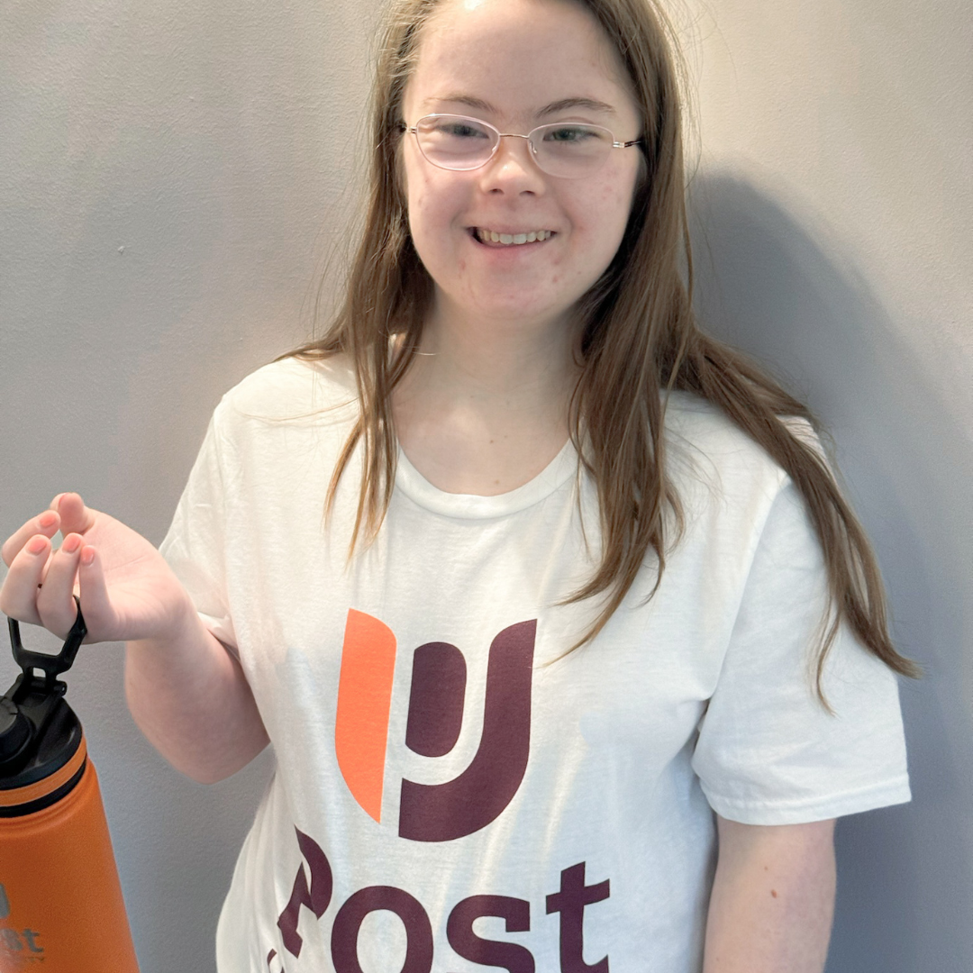Penny poses inside wearing a Post University t-shirt and holding a Post University orange water bottle as she smiles at the camera