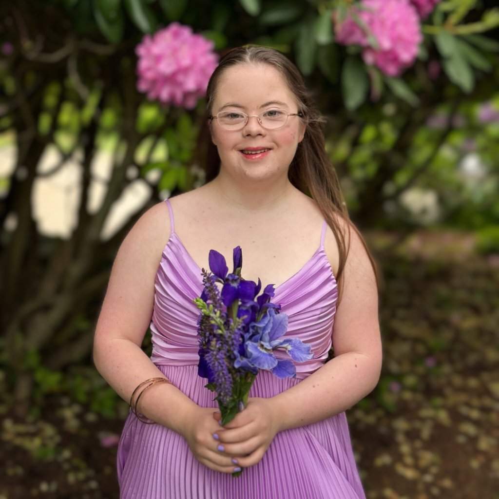 Penny smiles for the camera in front of a flowering bush. She is wearing her purple prom dress and holding dark purple flowers.